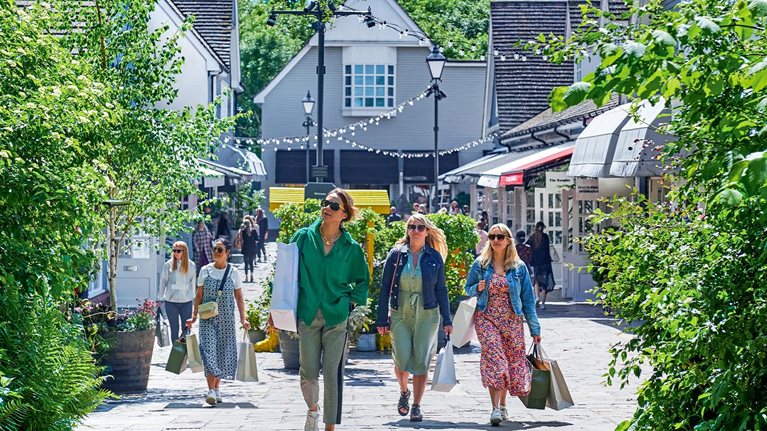Shoppers carrying shopping bags walk outdoors through Bicester Village. 