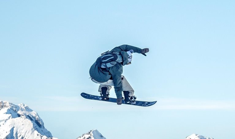 A snowboarder is captured midair performing a jump against a clear blue sky, with rugged, snow-covered mountains stretching across the background. The scene conveys motion and adventure, highlighting the contrast between the airborne rider and the vast alpine landscape below.