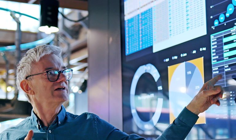 A man gesturing towards a large screen displaying graphs and data as he leads a presentation.