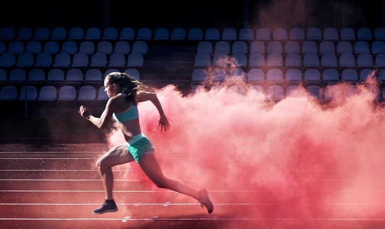 A determined female athlete races along a track, with a vibrant cloud of red smoke trailing behind her.