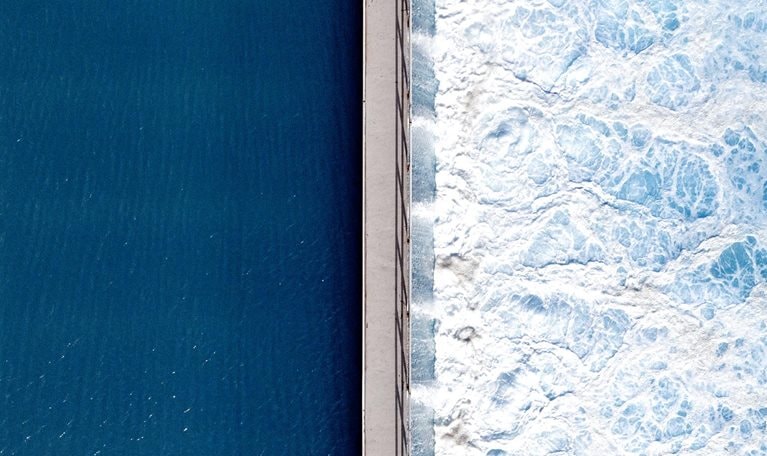 An overhead shot captures the stark contrast between deep, tranquil blue water on one side and churning, white-foamed turquoise waves on the other, bisected by a narrow concrete pier. The smooth, dark surface on the left stands in dramatic opposition to the textured, frothy turbulence crashing against the right side of the man-made barrier.