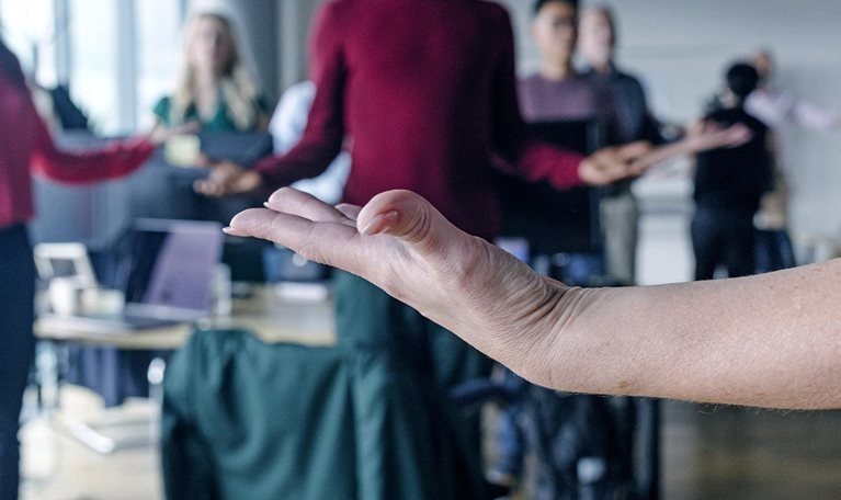 Close-up of a woman's upward facing hand in the foreground, with a blurred group of coworkers meditating in the background.