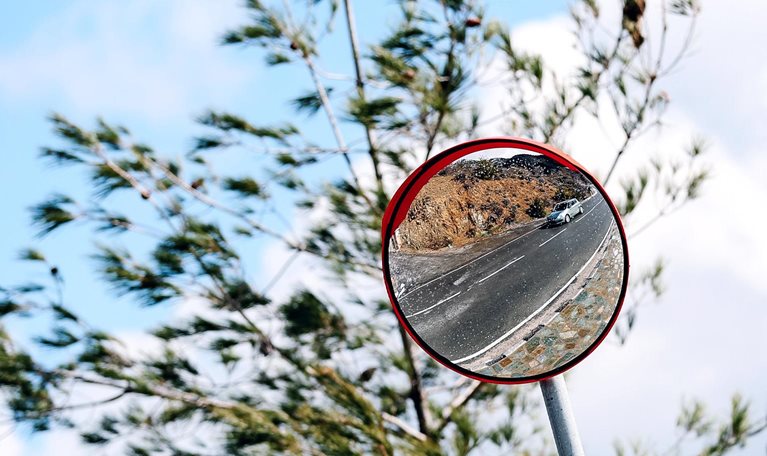 A round roadside convex mirror mounted on a pole, reflecting a winding road with a car driving along it. Behind the mirror, tree branches sway against a bright blue sky with scattered clouds.