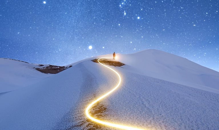 A solitary figure stands upon a snow-covered peak against a starry night sky. A zig-zagging light trail marks the path taken by the figure to reach the summit.