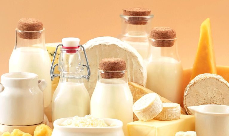 Assortment of dairy products including milk bottles, cheese varieties, butter, and cream on a light background.