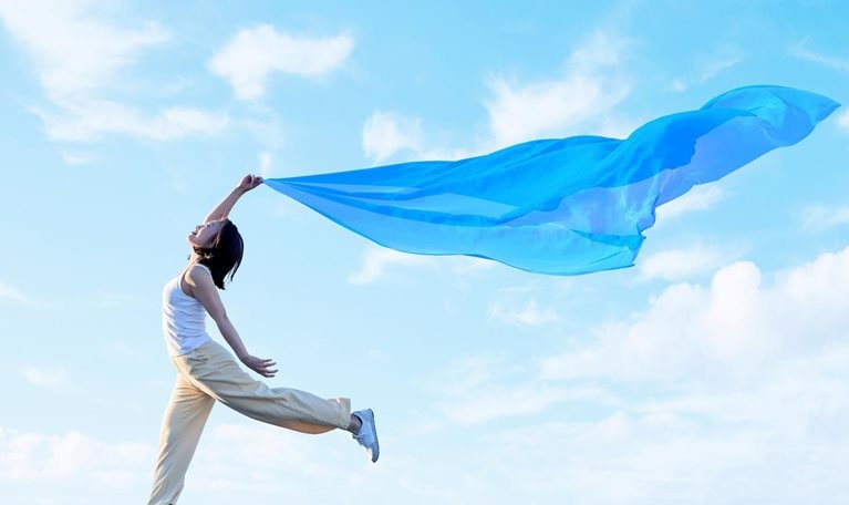 A young woman joyfully leaps through the air, holding a flowing blue scarf that billows dramatically against a bright blue sky with soft clouds.