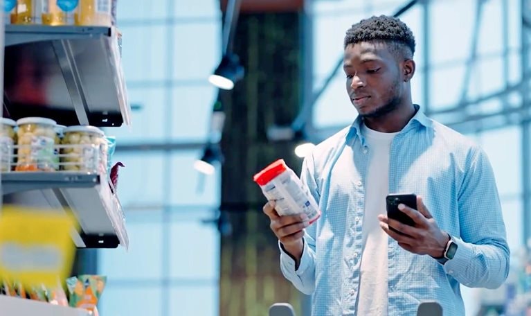 Person in a grocery store comparing a product label while holding a smartphone next to a shopping cart.