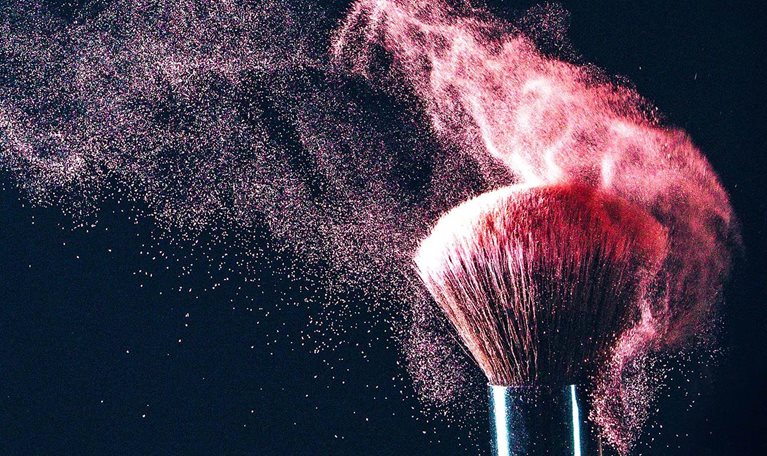 A makeup brush with blush particles artistically being shaken off