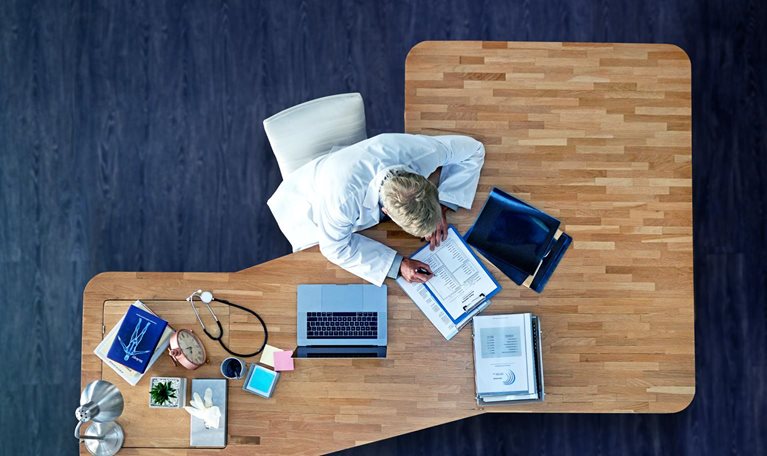 A bird's eye perspective of a physician sitting at an executive desk, filling out paperwork. Beside them sits a laptop, medical records, and a stethoscope. The desk is positioned on a rich, dark wooden floor.