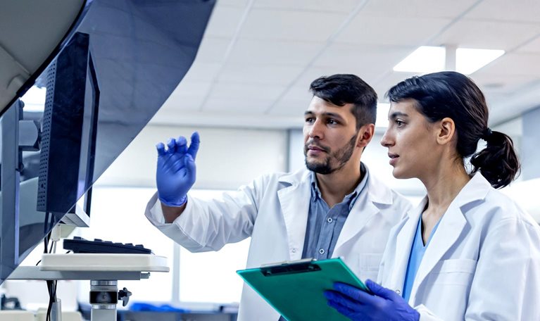 Two laboratory scientists in white coats and blue gloves stand side by side examining a large machine with a monitor. One scientist points to the screen while the other holds a clipboard, both appearing focused on the data or results displayed.