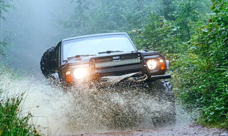 The image shows a rugged off-road vehicle driving through a muddy forest trail, splashing water as it moves forward. Dense green foliage surrounds the path, and mist hangs in the air, creating a dramatic and adventurous atmosphere.