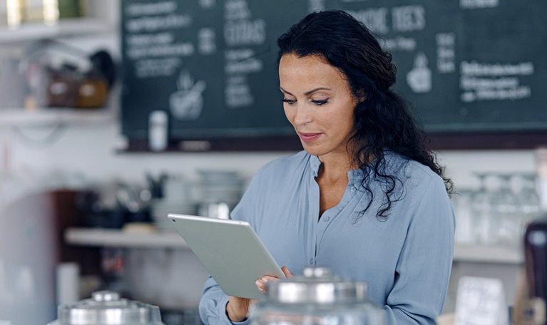 Brunette woman using digital tablet while standing at counter in coffee shop with the menu behind her.