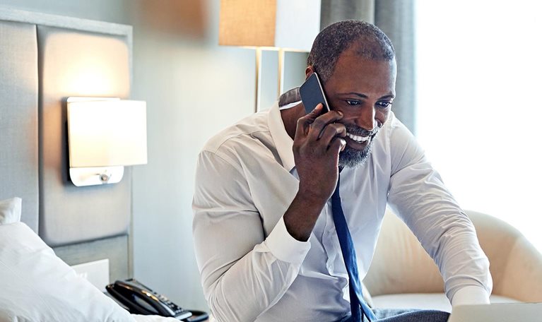 Male entrepreneur sitting on bed in a hotel room working on this laptop while on his cellphone