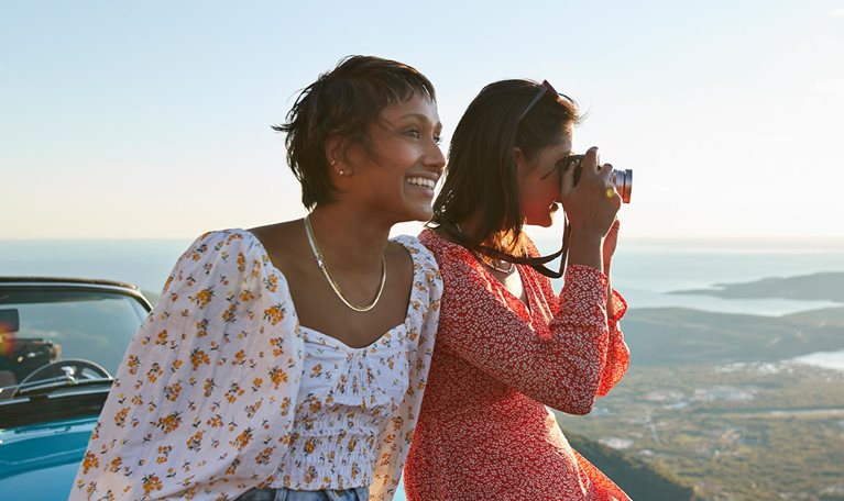 Close up image of friends on vacation sitting on the hood of a car smiling and taking pictures of the view in Montenegro.