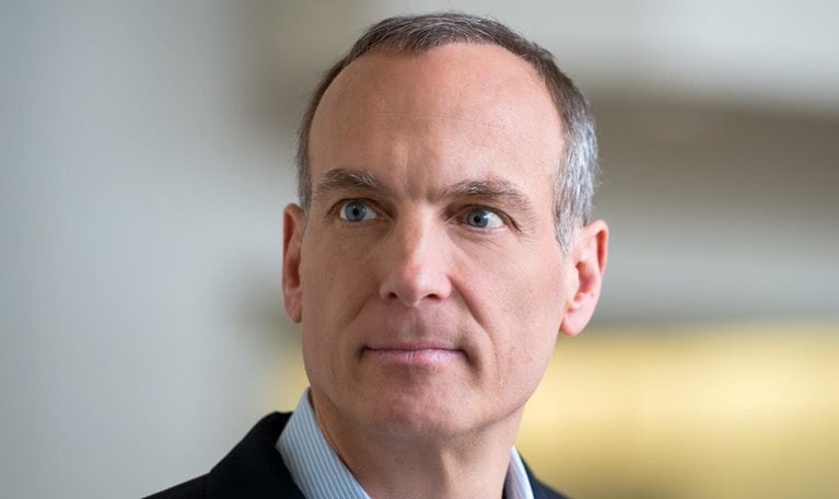 Glenn Fogel with short salt and pepper hair and blue eyes, wearing a dark blazer over a light collared shirt. He is looking slightly off to the side with a neutral, thoughtful expression against a softly blurred indoor background.