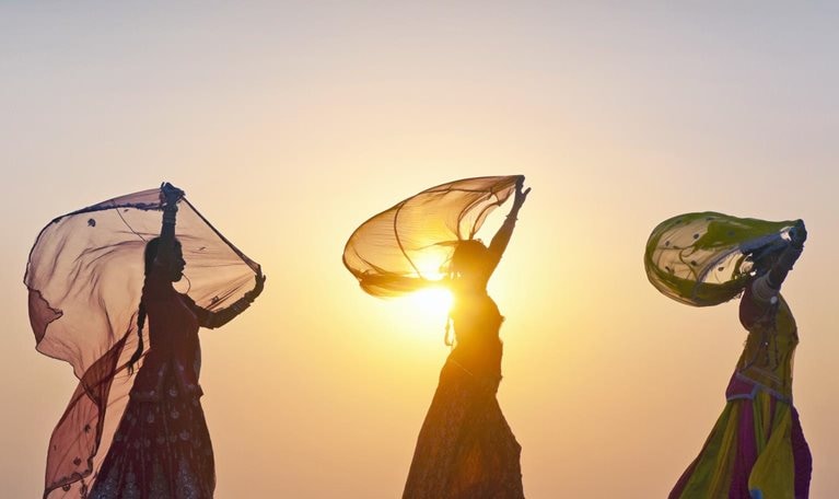 As the sun rises behind them, four Indian women dressed in traditional attire stroll along the crest of a desert dune, casting their figures in silhouette.