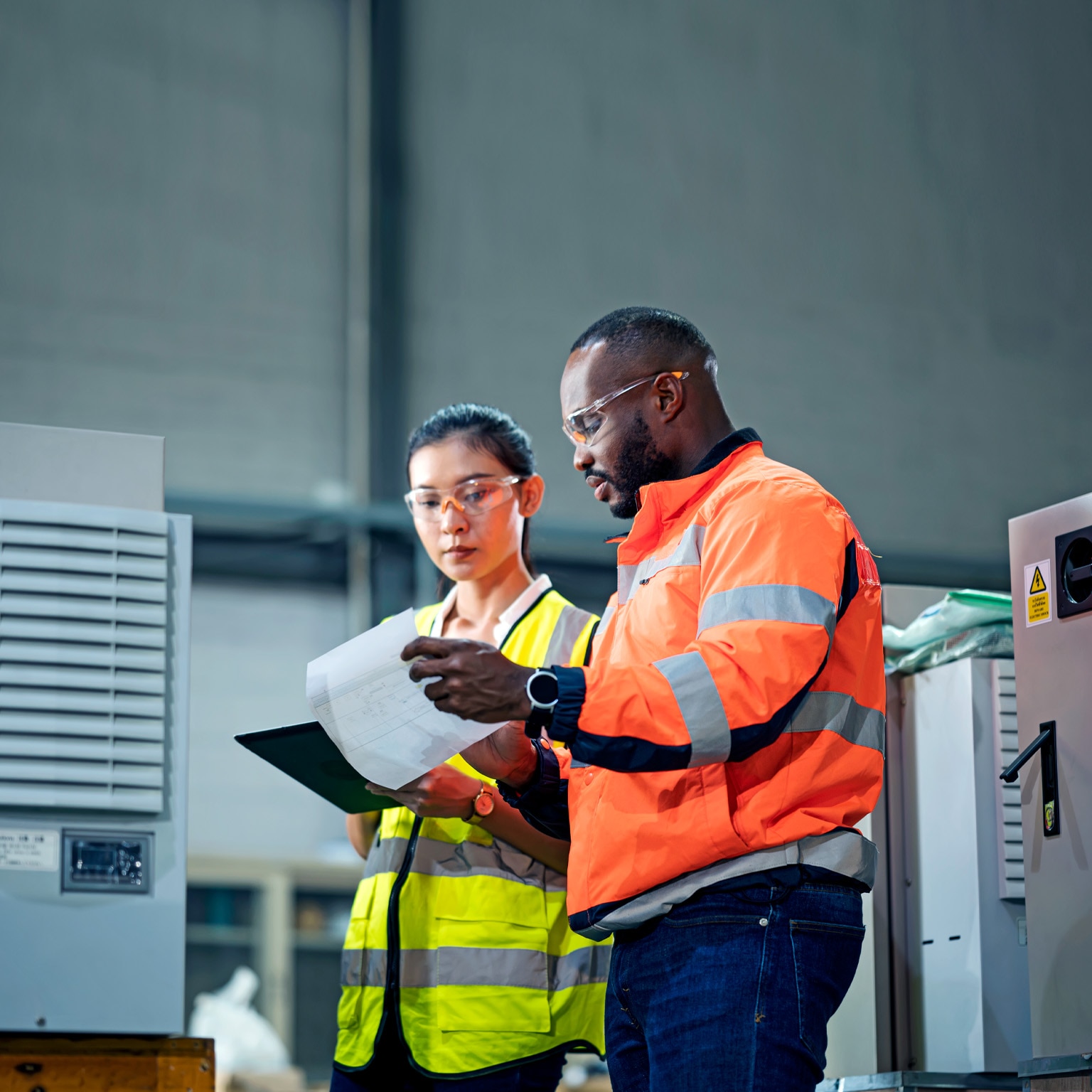 Image of a woman holding a tablet and a man with a checklist, both wearing different color-coded work jackets standing in a control room