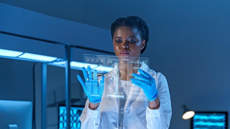 Image of a serious female scientist in a lab coat and protective gloves stands in a modern lab, working on a small HUD or graphic display