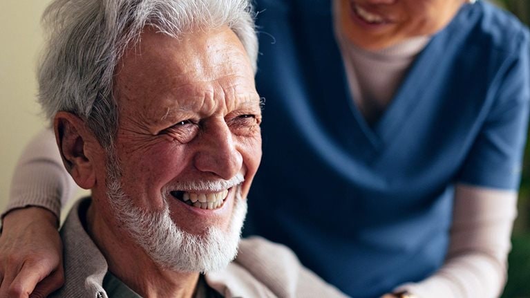 Portrait of a Smiling Elderly Man Taking a Jacket off With a Help of a Woman in a Care Home