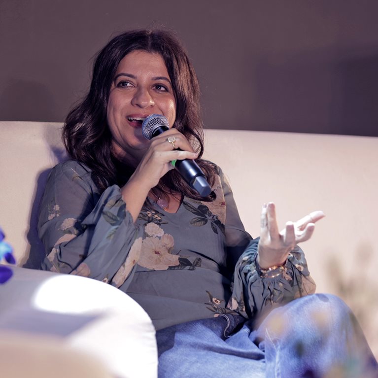 Zoya Akhtar seated on a white armchair speaks into a handheld microphone during a panel discussion. She gestures with one hand while smiling, wearing a floral blouse and jeans, against a softly lit background.