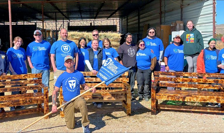 The Mission Continues’s volunteers posing outdoors behind newly built wooden benches, with one person kneeling in front holding a flag.
