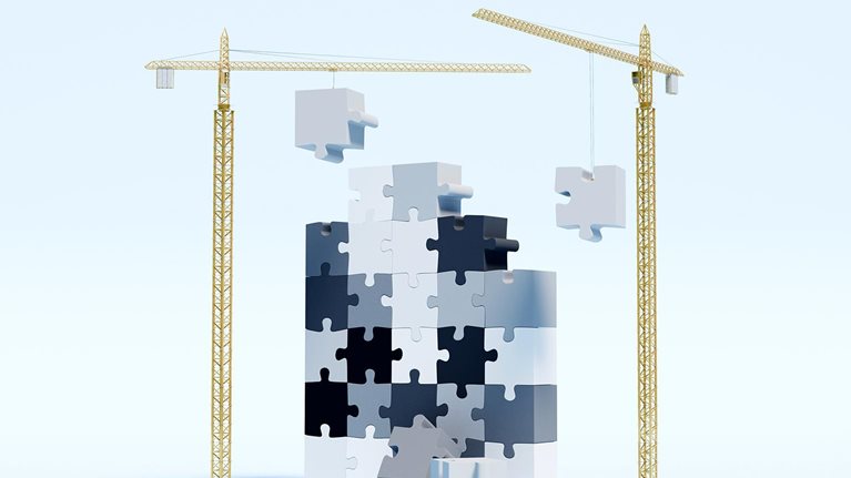 Construction cranes are actively lifting and placing puzzle pieces, slowly constructing a towering, upright puzzle.