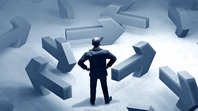 A multitude of large, concrete arrows pointing in different directions surround a businessman.