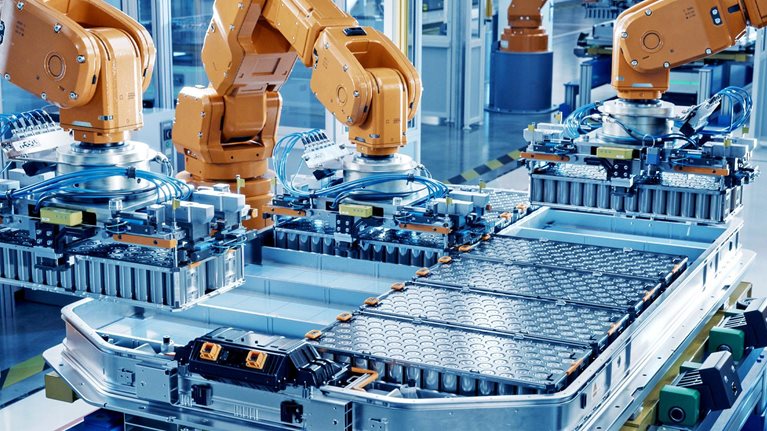 A modern factory floor where multiple orange industrial robotic arms are assembling large battery packs on an automated production line. The environment is clean, brightly lit, and highly organized, emphasizing advanced manufacturing and automation technology.