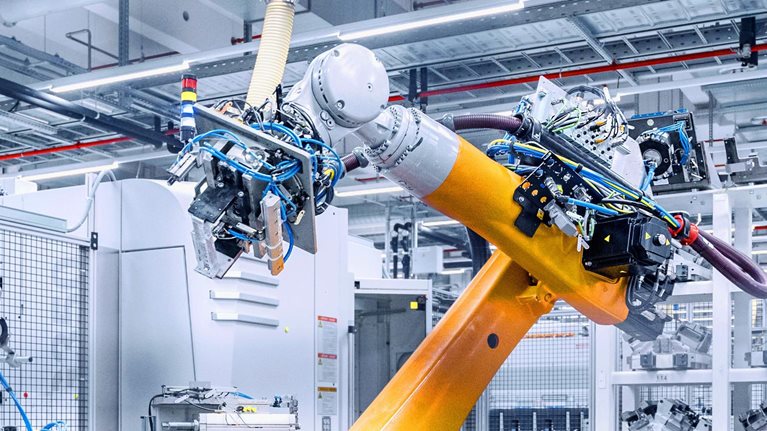 A large, bright industrial workspace with a prominent yellow robotic arm in the foreground. The robot appears to be working within an automated manufacturing system, surrounded by other machinery, parts, and storage racks.