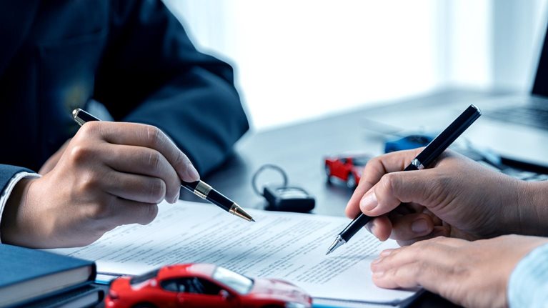 Two people signing documents at a desk, with their hands holding pens over a contract. Toy cars and a set of keys on the table suggest the paperwork is related to an auto insurance agreement.