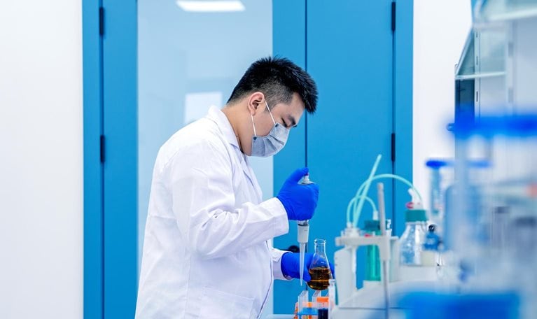 A lab researcher wearing a face mask, gloves, and a white coat is using a pipette to transfer liquid into a flask on a laboratory bench. The scene shows a clean, modern lab environment with scientific equipment and containers arranged for experimental work.