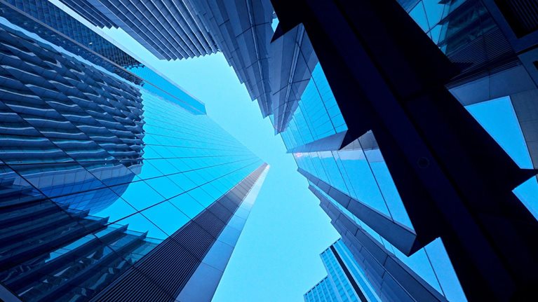 A dramatic upward view of modern glass skyscrapers converging toward the sky. The sharp lines and reflective surfaces create a sleek, futuristic feel.