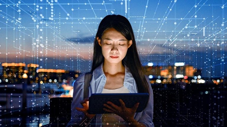 A woman stands outdoors at dusk, focused on a tablet in her hands with a city skyline glowing in the background. Digital lines and data-like patterns overlay the scene, suggesting connectivity, technology, and data flow.