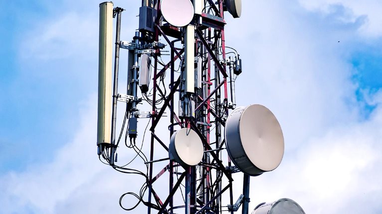 A tall telecommunications tower rises against a partly cloudy blue sky, covered with multiple circular and rectangular antennas.