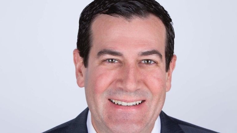 Portrait of Mark Birkhead with a light smile, wearing a charcoal suit and a maroon tie.