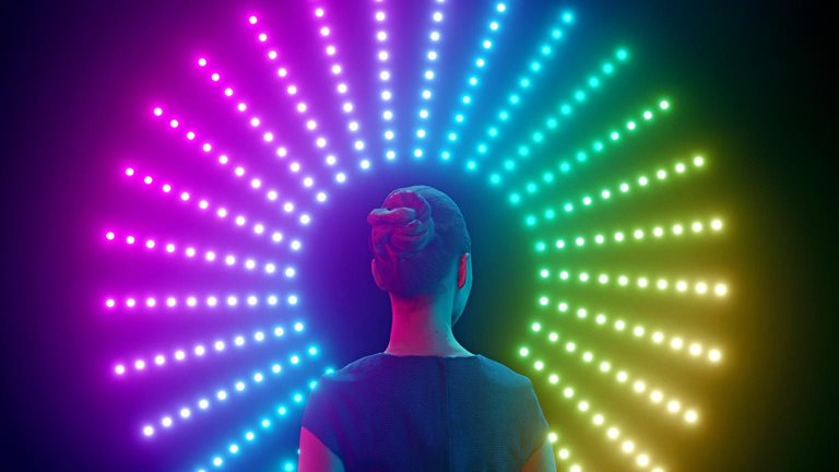 A woman with her back to the viewer stands in front of a circular arrangement of colorful lights. The lights are arranged in a rainbow pattern, transitioning from pink to blue to yellow.