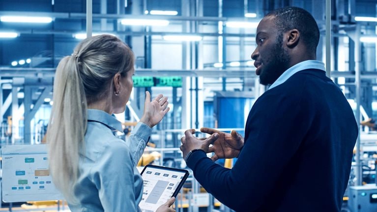 Two engineers stand in a high-tech automotive manufacturing facility discussing data displayed on digital screens and a tablet, with robotic machinery operating in the background.