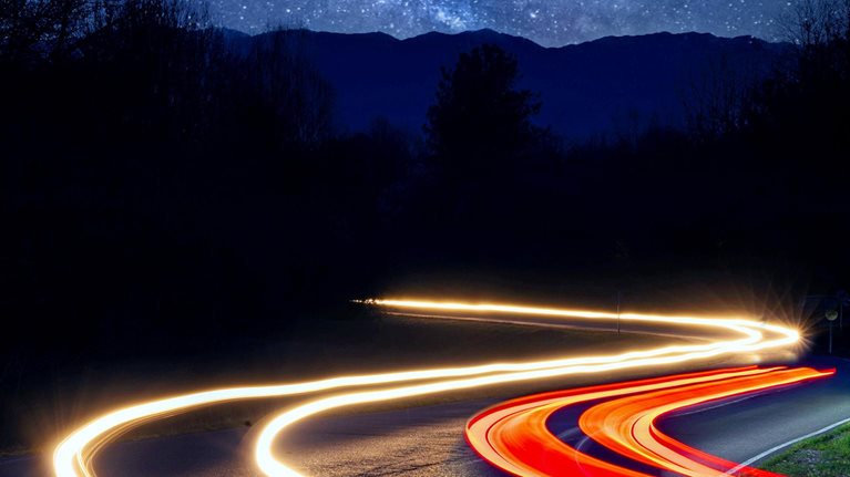 car headlight trails driving on a road through a forest on a starry night with the milky way and mountains in the background