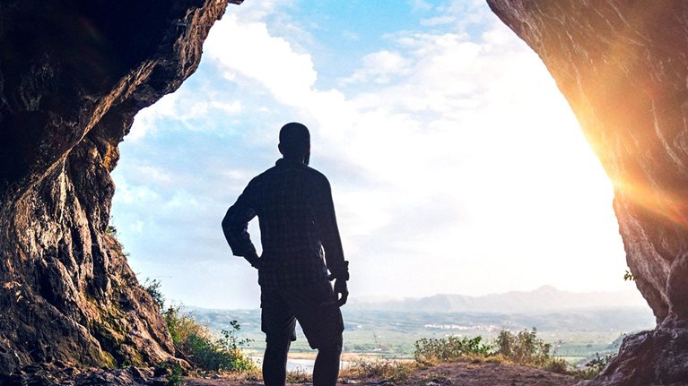 A hiker looks out over a sunset from inside the large opening of a cave