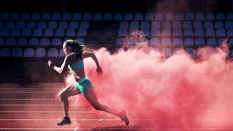 A determined female athlete races along a track, with a vibrant cloud of red smoke trailing behind her.