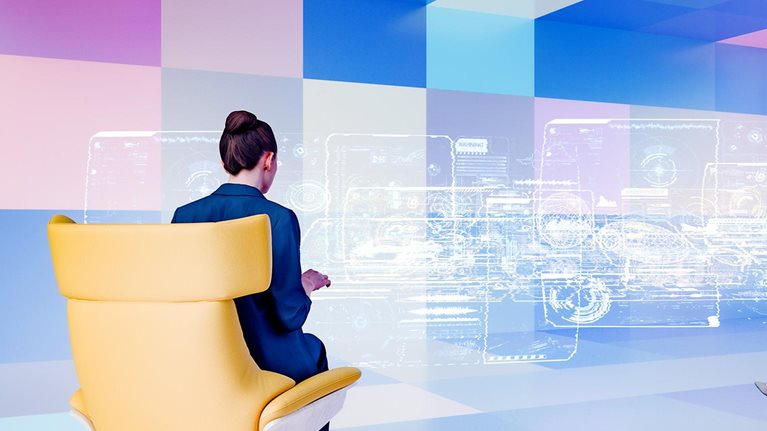 Two businesswomen sit in modern chairs facing a large, translucent screen displaying complex data visualizations. The futuristic setting suggests a collaborative work environment utilizing advanced technology.