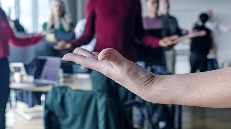Close-up of a woman's upward facing hand in the foreground, with a blurred group of coworkers meditating in the background.