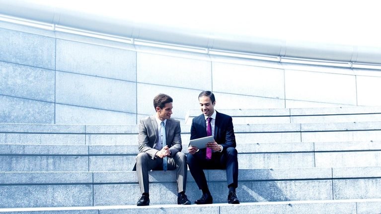 A manager giving feedback to an employee. They are sitting on a set of stairs while looking at a tablet.