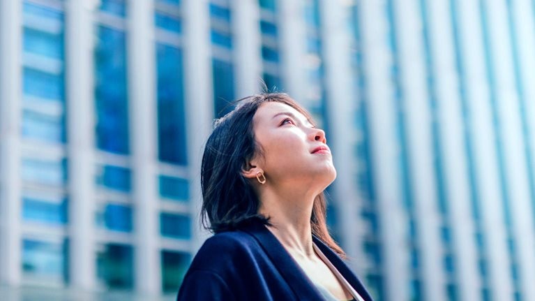 A modern business district with tall buildings is the background for a young, confident businesswoman looking up at the sky from a low angle.