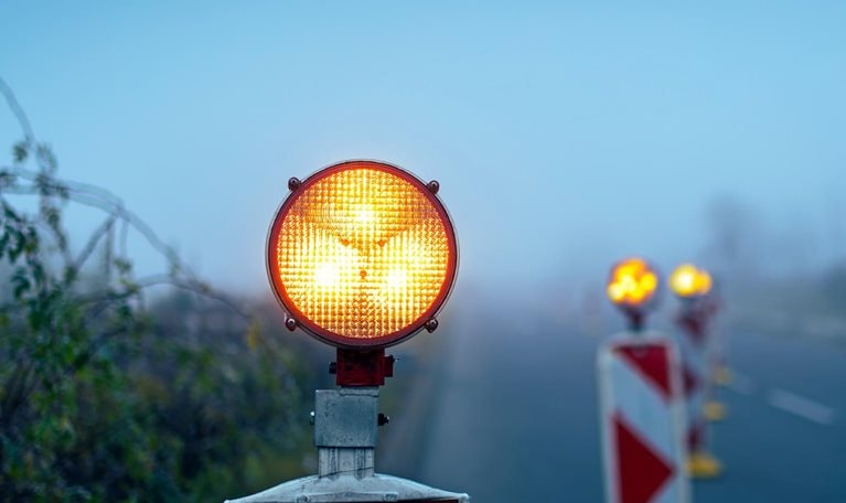 A bright amber warning light sits atop a road barrier, glowing vividly against a foggy, muted background. Additional blurred warning lights and striped barriers line the roadside.