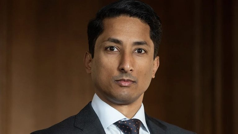 Jean-Paul Carvalho wearing a dark suit, white shirt, and patterned tie looks directly at the camera with a neutral, composed expression. He stands against a warm-toned, softly blurred background.