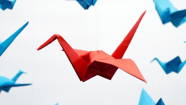 One bold red origami crane hangs from a string among a plethora of blue origami cranes also suspended in the air.