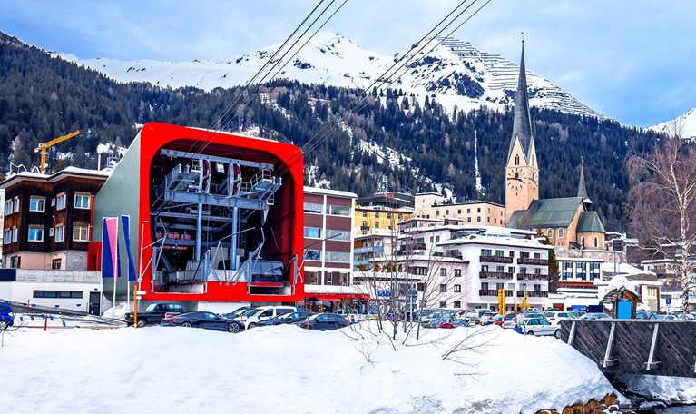 From Davos, Switzerland, one can see the tramway that takes you to the ski resort. Alongside the tram, there are small buildings, while the background is dominated by mountains. A stream flows in the foreground, adding to the overall view.