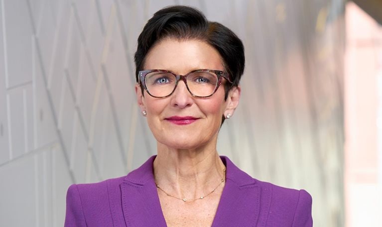 A portrait of Jane Fraser with short dark hair wearing glasses and a purple blazer over a black top. She stands against a softly blurred, modern interior background and looks directly at the camera with a confident, composed expression.