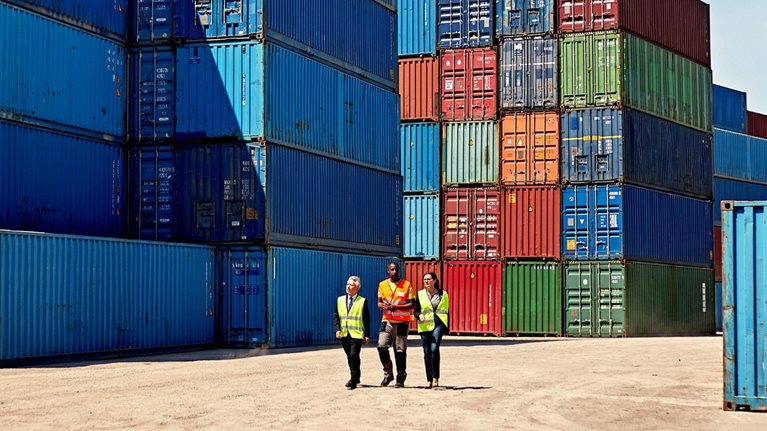 Three people in high-visibility vests walking in a shipping yard, amidst stacks of colorful shipping containers. The scene suggests a meeting in a bustling port or logistics area.